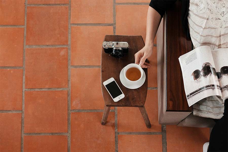 A girl is holding cup of tea on the table and there is phone and camera and reading book